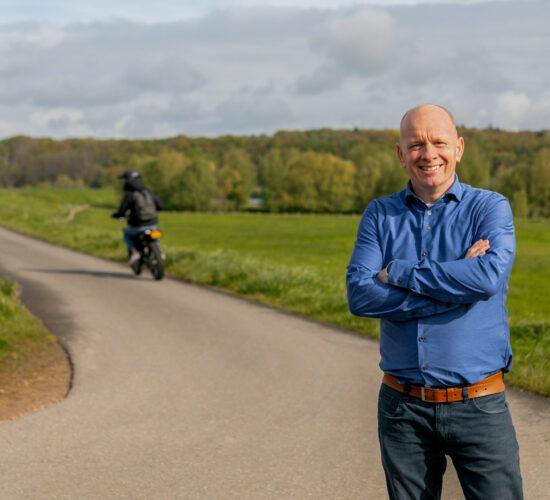 Collega Arie lacht naar de camera. Achter hem zie je een brommerrijder.