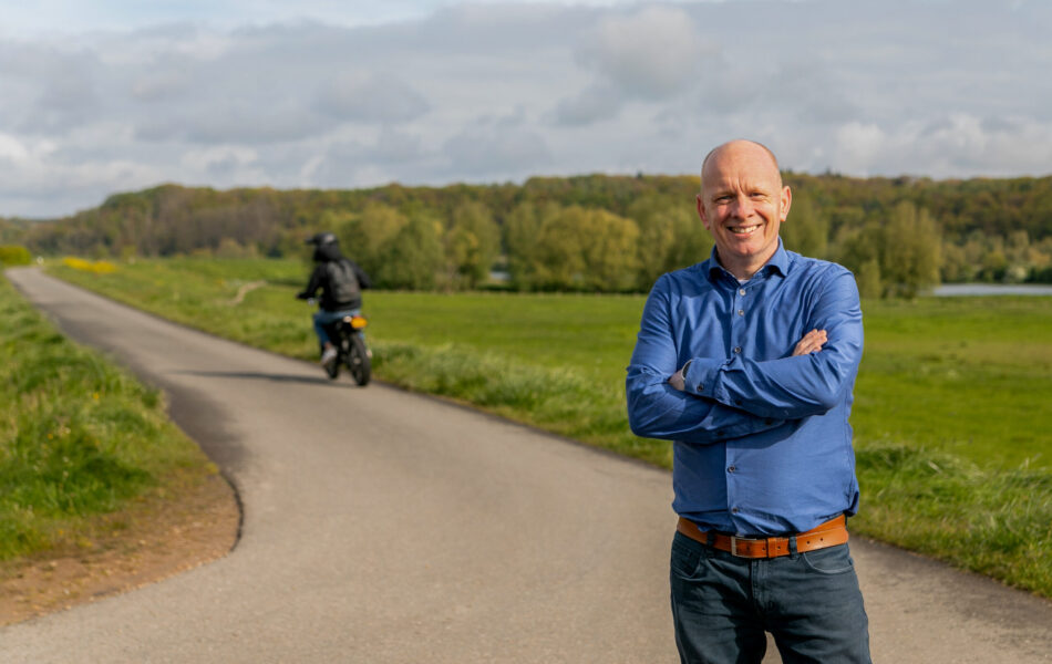 Collega Arie lacht naar de camera. Achter hem zie je een brommerrijder.