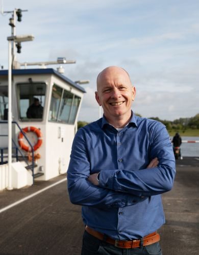 Collega Arie lacht naar de camera. Achter hem zie je de veerpont van Opheusden.